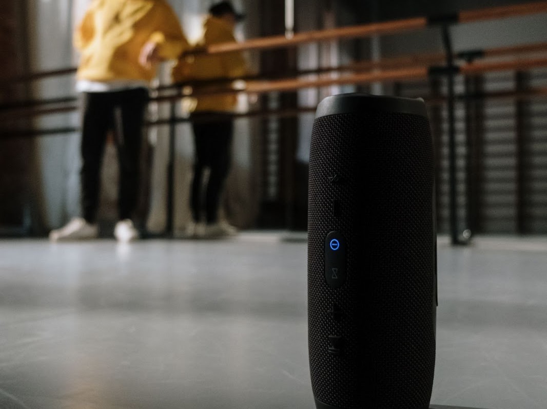 Aparelho de som sem fio portátil tocando música via bluetooth em uma academia de dança, ao fundo é possível ver um rapaz se preparando para dançar.