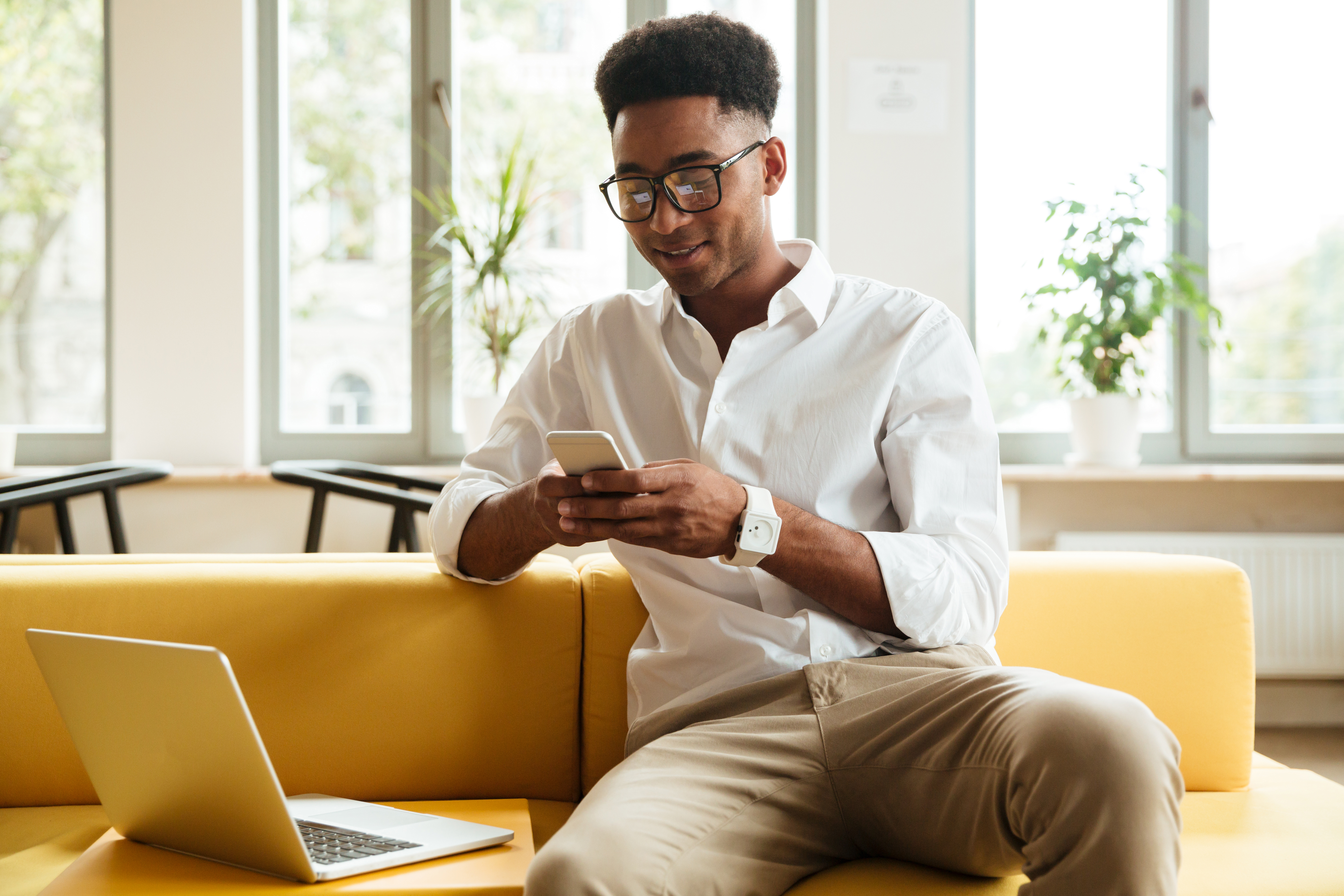 Na imagem um belo homem afrodescendente de camisa branca e óculos de grau, está sentado em um sofá amarelo em frente a um notebook enquanto conversa cm alguém via whatsapp.