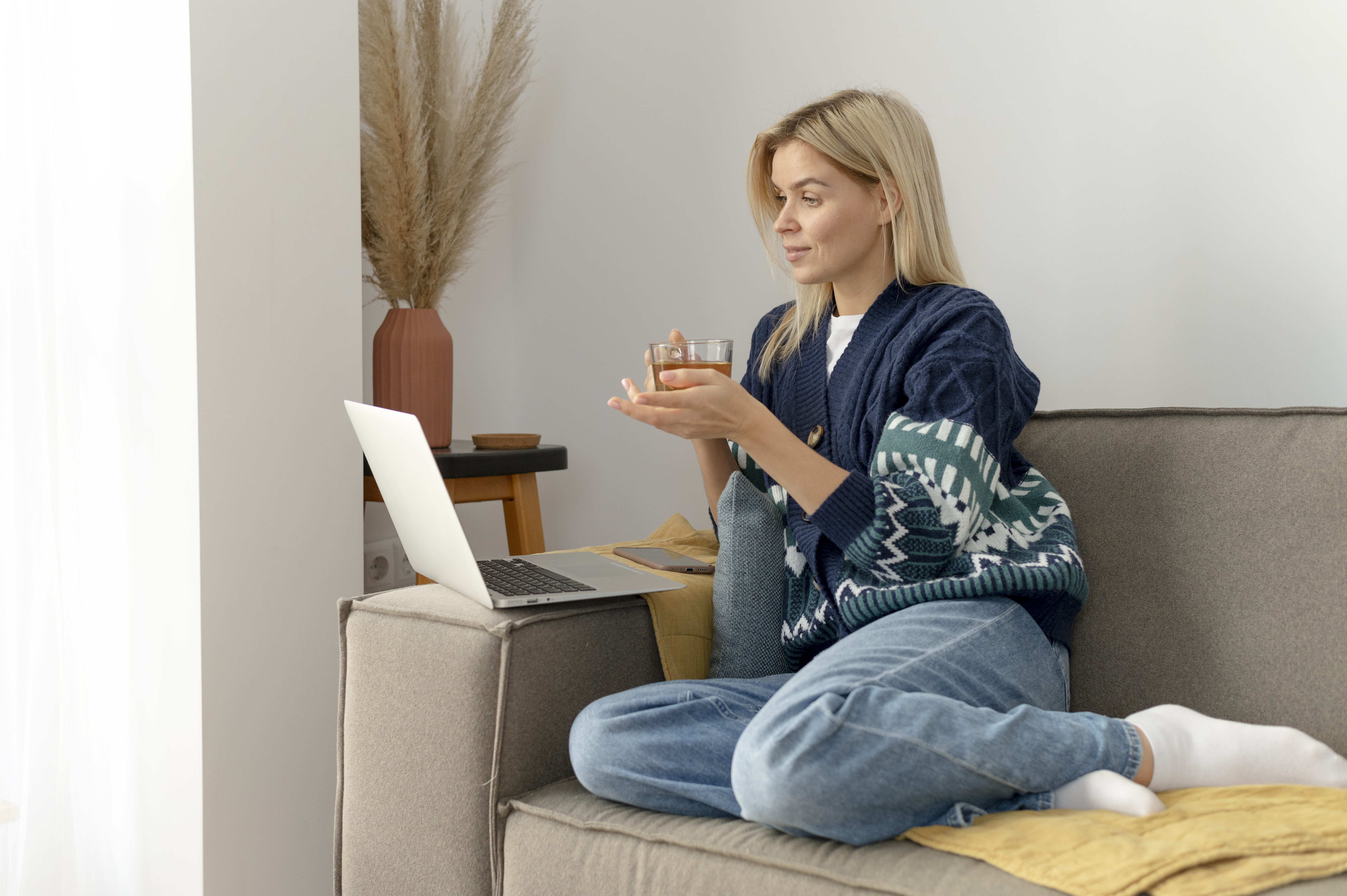 Uma mulher loira está sentada em um sofá e segurando uma xícara de chá enquanto olha um notebook aberto.